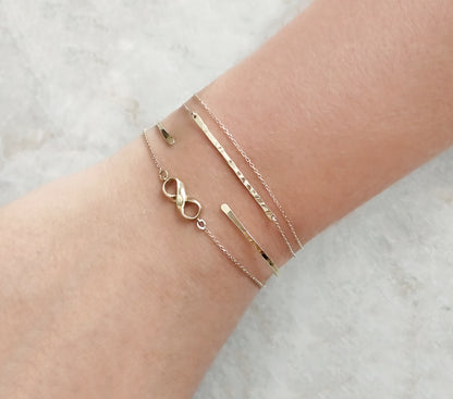 female wrist wearing a bracelet stack with a gold thin hammered bar bracelet with double chain, a thin hammered open bangle bracelet and an infinity charm bracelet