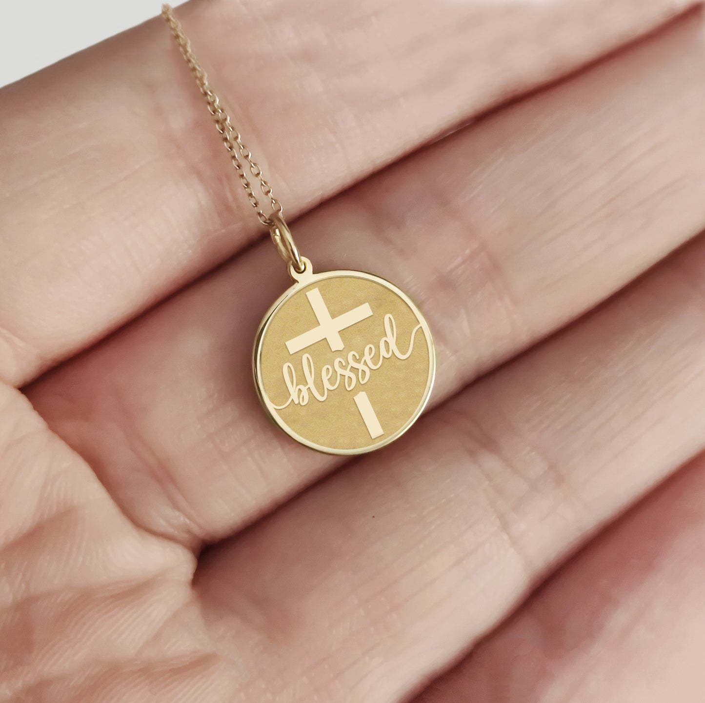 hand holding a gold necklace with a disc pendant engraved with the word blessed and a cross 