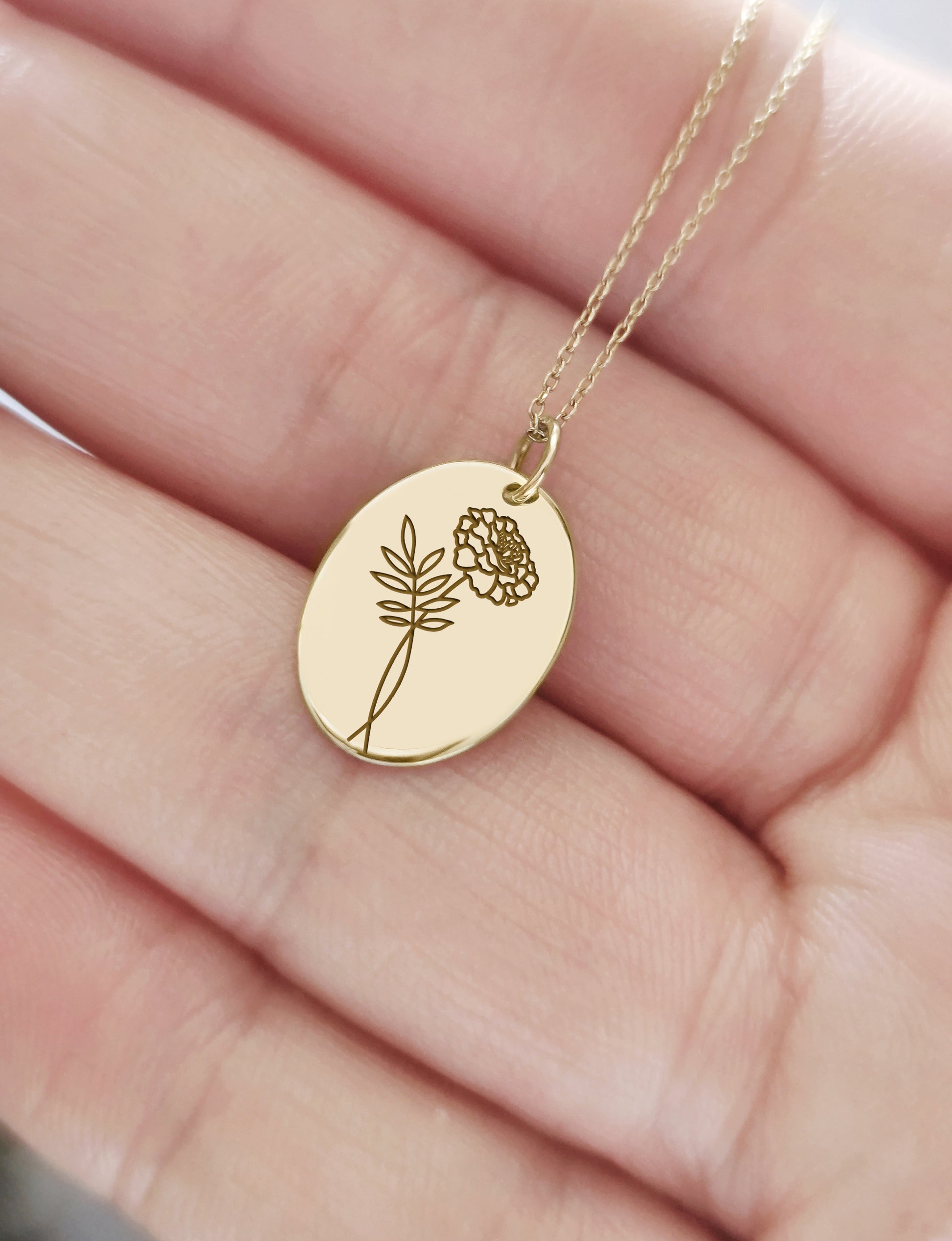 hand holding gold oval pendant necklace engraved with a Marigold the birth flower of October for size reference